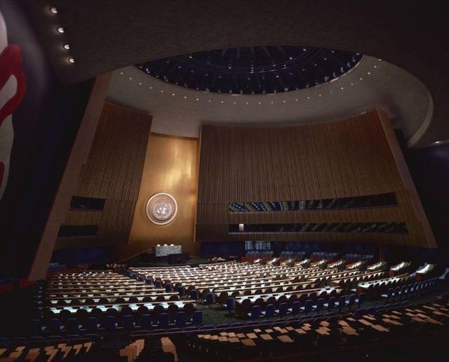 The United Nations General Assembly hall, empty, with the UN emblem above the rostrum.