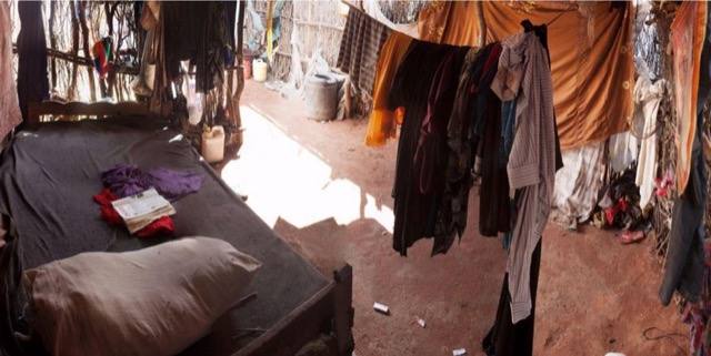 Interior of a refugee camp dwelling: a bed with a pillow and clothes hanging from lines.