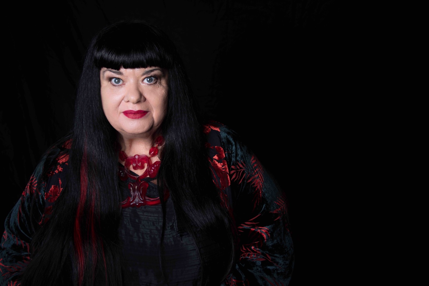 Lynette Wallworth, in a dark velvet jacket patterned with red flowers and a carved red necklace, photographed against a black background.